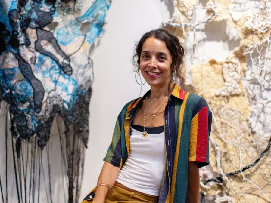 A woman seated on a wooden bench in front of two large hanging textile artworks