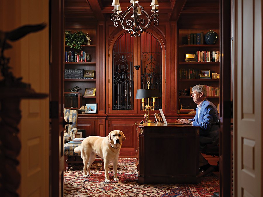 A man seated at a desk with a laptop, a yellow Labrador beside him